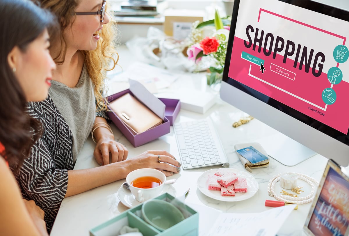 Two women shopping online on desktop computer browsing eCommerce store with credit card and products on desk.